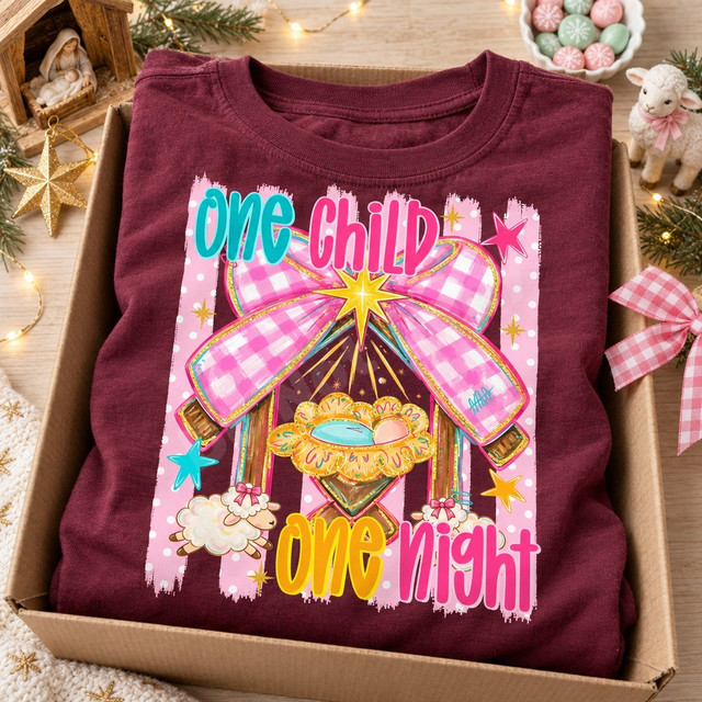 One Child One Night Christmas DTF transfer with baby Jesus in a golden manger, pink gingham bow, gold star, and lambs on a pink polka-dot striped background Shown on a Wine t-shirt.