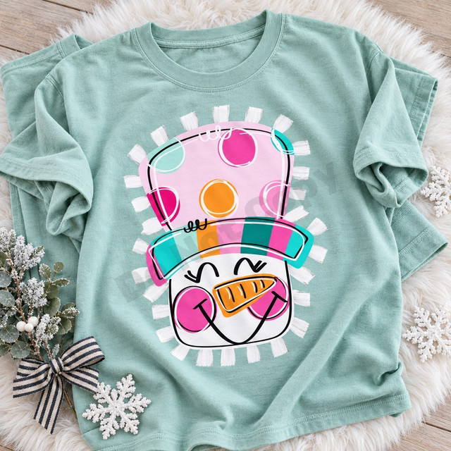 Colorful snowman face DTF transfer with polka-dot striped hat, carrot nose, pink cheeks, and winking eyes framed by a fringed border Shown on a Seafoam t-shirt.
