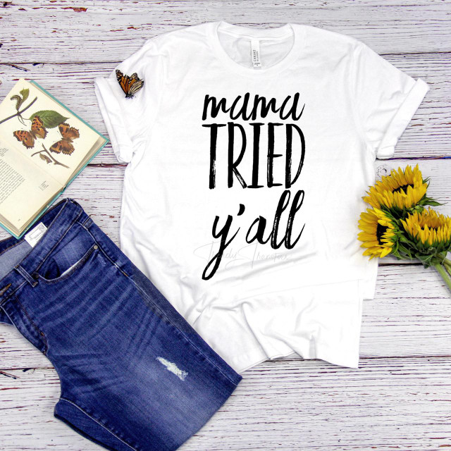 Mama Tried Y'all funny Sublimation Transfer, white T-shirt with black handwritten-style text, displayed on wooden background with sunflower bouquet, jeans, and butterfly book.