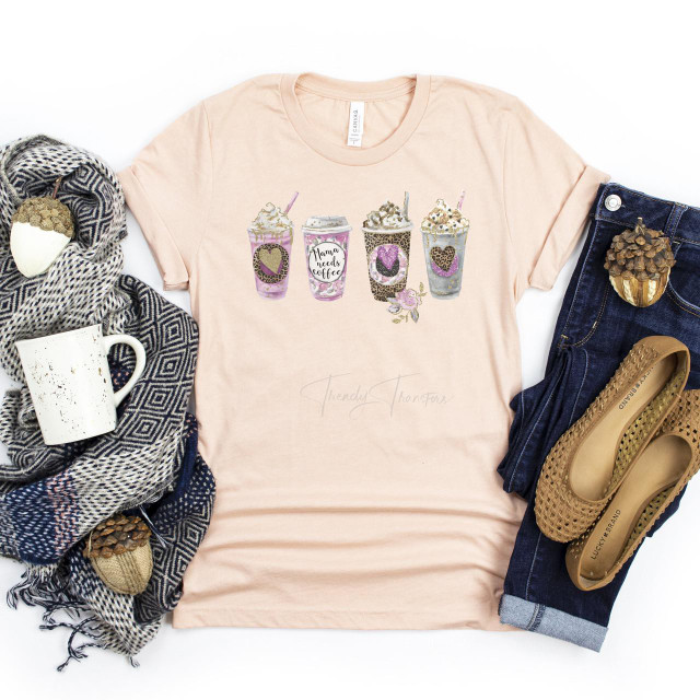 Mama Needs Coffee Watercolor Screen Print Heat Transfer, a pastel pink T-shirt with watercolor coffee cups design, surrounded by a mug, scarves, jeans, shoes, and decorative acorns.