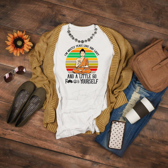 Peace Love Light Sublimation Transfer T-shirt with Buddha graphic, colorful stripes, and humorous quote, displayed with casual accessories on a wooden surface.