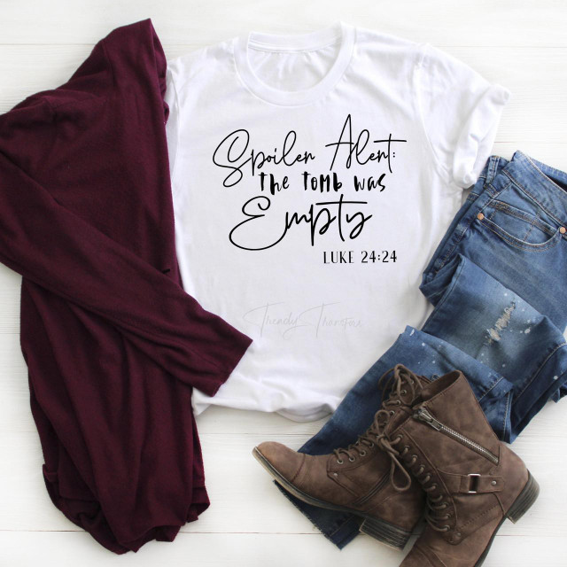 Spoiler Alert: The tomb was empty Easter Luke 24:24 Bible verse religious Sublimation Transfer T-shirt, white t-shirt with black handwritten style text and Bible verse, paired with jeans, brown boots, and a maroon scarf on a white wooden background.