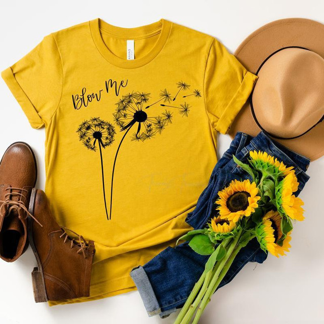 Product Blow Me Dandelion Screen Print Transfer, yellow T-shirt with dandelion flower graphic and text, alongside a pair of brown boots, a tan hat, and a bunch of sunflowers on blue jeans.