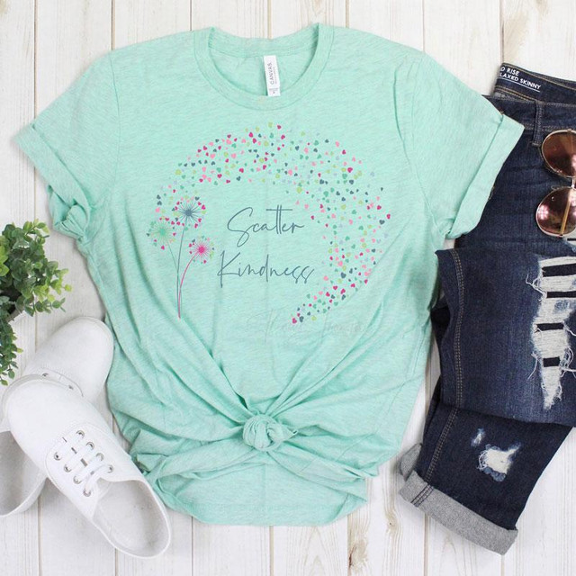 Scatter Kindness Heart Screen Print Heat Transfer, a light green T-shirt with a colorful heart and dandelion design, laid flat with distressed jeans, white sneakers, and sunglasses.