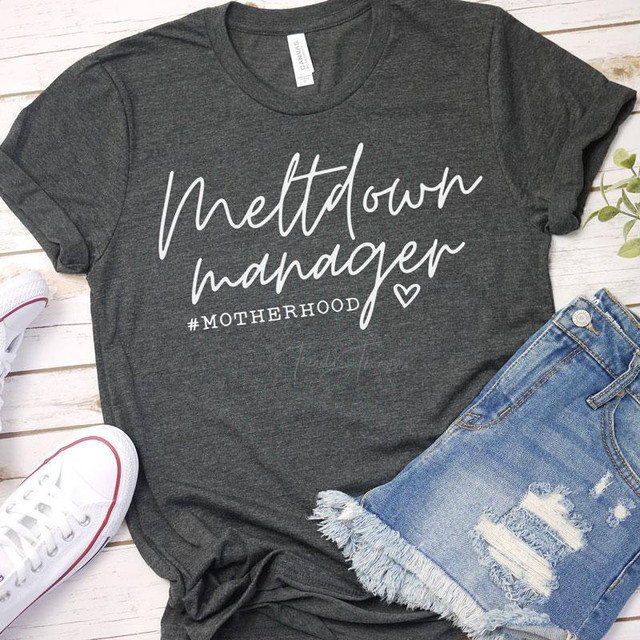 Meltdown Manager Motherhood Screen Print Transfer, a casual dark gray T-shirt with white handwritten-style text and hashtag, displayed with denim shorts and sneakers on a wooden background.