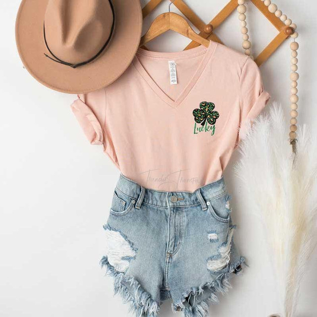 Lucky Shamrock HAT/POCKET Screen Print Heat Transfer, pink t-shirt with green shamrock design and Lucky text, distressed denim shorts, beige hat hanging on a wooden hanger.