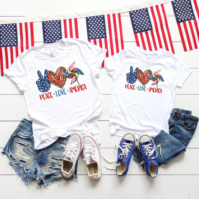 Peace Love America Pinwheel red white and blue flag Patriotic Sublimation Transfer, patriotic t-shirts with American flag-themed pinwheel design and peace sign, paired with denim shorts and sneakers, on a white wooden background with American flags.