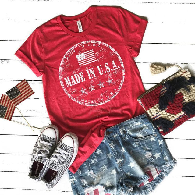 Made In The USA WHITE Screen Print Heat Transfer, close-up view of a fashionable t-shirt with a patriotic American flag design and distressed denim shorts, on a white wooden background with American flags and a patriotic woven clutch.