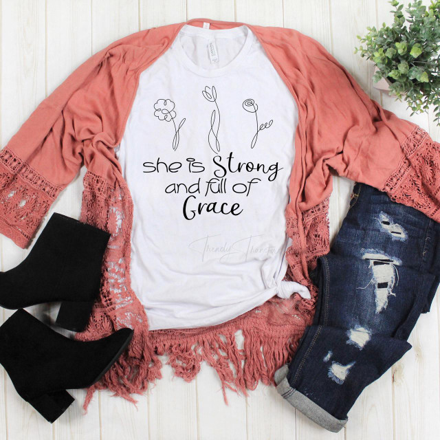 She is strong and full of grace floral sublimation transfer, white t-shirt with floral line art and inspirational quote, styled with distressed jeans, a pink lace-trimmed cardigan, and black ankle boots, arranged on a white wooden surface.