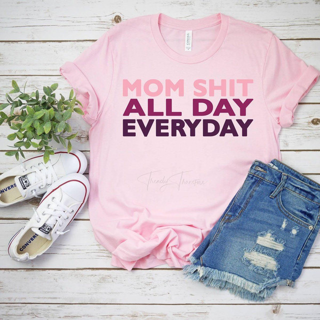 Mom Shit All Day Every Day FINAL STOCK Screen Print Heat Transfer T-shirt, pink casual short sleeve tee with bold text, laid out on white wooden background with sneakers, denim shorts, and a green plant.