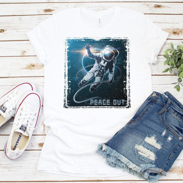 Peace Out Sublimation Transfer, space-themed astronaut design printed on white T-shirt, laid on wooden surface with denim shorts and white sneakers beside it.