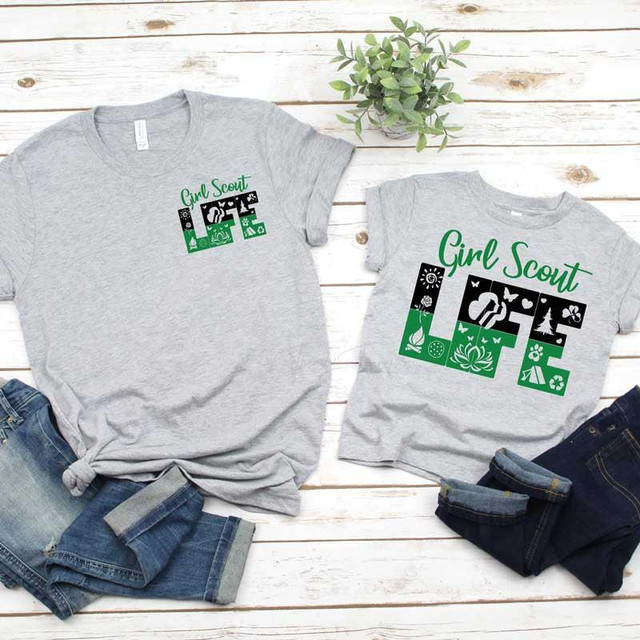 Girl Scout Life Youth Screen Print Transfer, two gray t-shirts with green and black Girl Scout themed designs, laid on a white wooden surface with denim jeans and a potted plant.