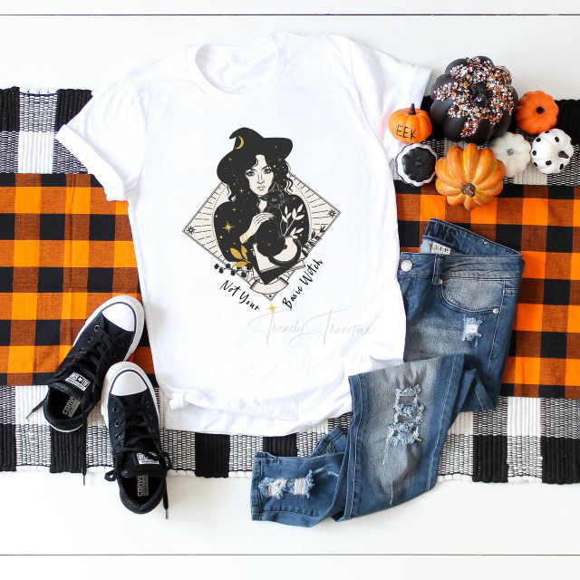 Not Your Basic Witch Sublimation Transfer-1655982304, a Halloween-themed white T-shirt with a black witch illustration and the phrase 'Not Your Basic Witch', styled with distressed jeans and Halloween pumpkins on a checkered tablecloth.
