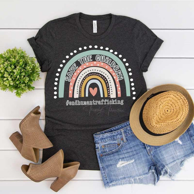 Save The Children Rainbow with White Outline Screen Print Heat Transfer, black t-shirt with rainbow design and hashtag #endhumantrafficking, alongside a straw hat, distressed denim shorts, beige ankle boots, and a green plant on a white wooden background.