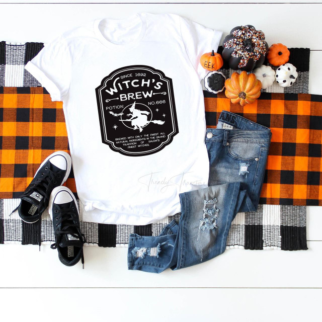 Witch's Brew Potion Sublimation Transfer, Halloween-themed black and white graphic design on a white T-shirt, displayed alongside pumpkins, a pair of black and white sneakers, and distressed denim jeans on a checkered orange and black tablecloth.
