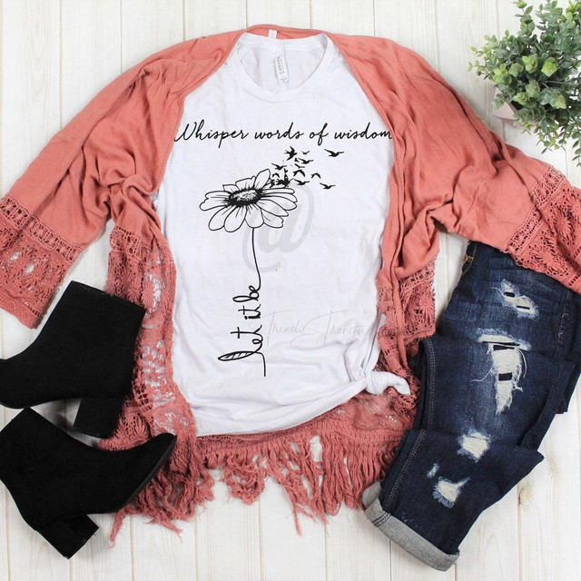 Whisper Words Of Wisdom Daisy Sublimation Transfer, white T-shirt with a daisy flower and flying birds design, paired with a peach lace cardigan, distressed jeans, and black ankle boots, laid out on a white wooden surface with a green potted plant.