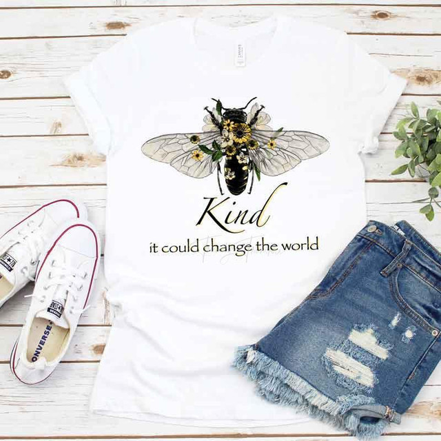 Be Kind It Could Change The World Sublimation Transfer T-shirt with bee and floral design, white casual tee, next to denim shorts and white sneakers, on wooden background