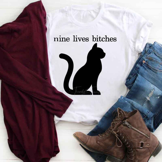 Nine Lives Bitches Sublimation Transfer T-shirt with a black cat silhouette and text, casual outfit laid out with jeans, brown boots, and a maroon cardigan.