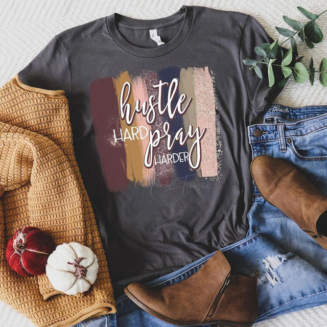 Hustle Hard Pray Harder Screen Print Heat Transfer T-shirt, dark gray, with motivational text design, accompanied by brown boots, denim jeans, a knitted beige sweater, and decorative pumpkins and greenery for styling.