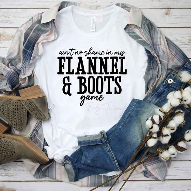 Ain't No Shame In My Flannel And Boots Game Sublimation Transfer, white t-shirt with black text, casual fashion style with plaid shirt, jeans, and cowboy boots, laid out on a wooden background with cotton branches for a rustic look.