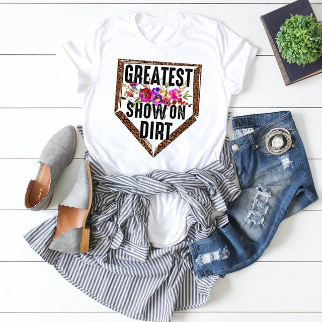 Greatest Show On Dirt Leopard Sublimation Transfer, white t-shirt with a leopard print border and colorful floral design, laid out with denim jeans, gray shoes, and a striped shirt on a white wooden surface with a black book and a green plant.