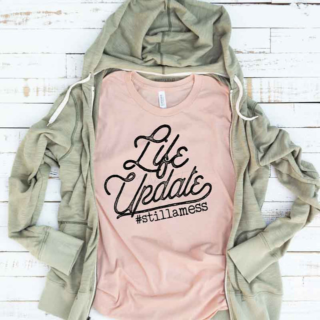 Life Update Still A Mess Print Heat Transfer, pink T-shirt with black text and hashtag, laid on a white wooden surface with a beige hoodie.