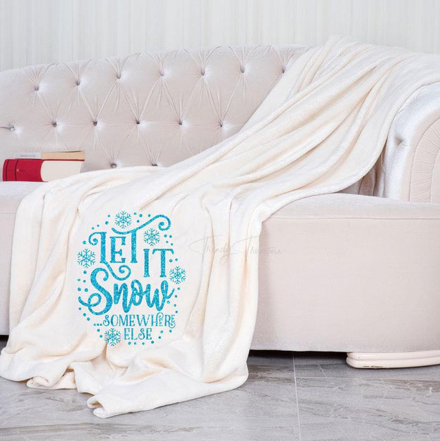 Let It Snow Somewhere Else Sublimation Transfer, image of a white blanket with a blue snowflake and text design on it draped over a beige tufted sofa.