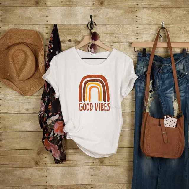Good Vibes Fall Rainbow Sublimation Transfer, white T-shirt with fall rainbow design and 'Good Vibes' text, hanging on a wooden wall among other fashion accessories.