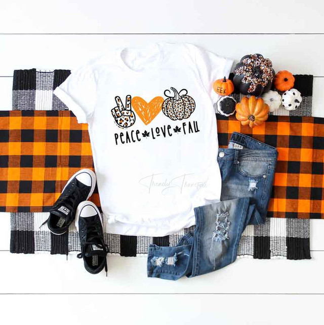 Peace Love Fall Sublimation Transfer, white T-shirt with fall-themed graphic and text, surrounded by autumn decorations and casual wear on a checkered tablecloth.