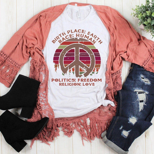 Birth Place Earth Hippie Sublimation Transfer, printed t-shirt with peace symbol and environmental message, with pink lace cardigan, distressed jeans, and black ankle boots laid out on a wooden surface.