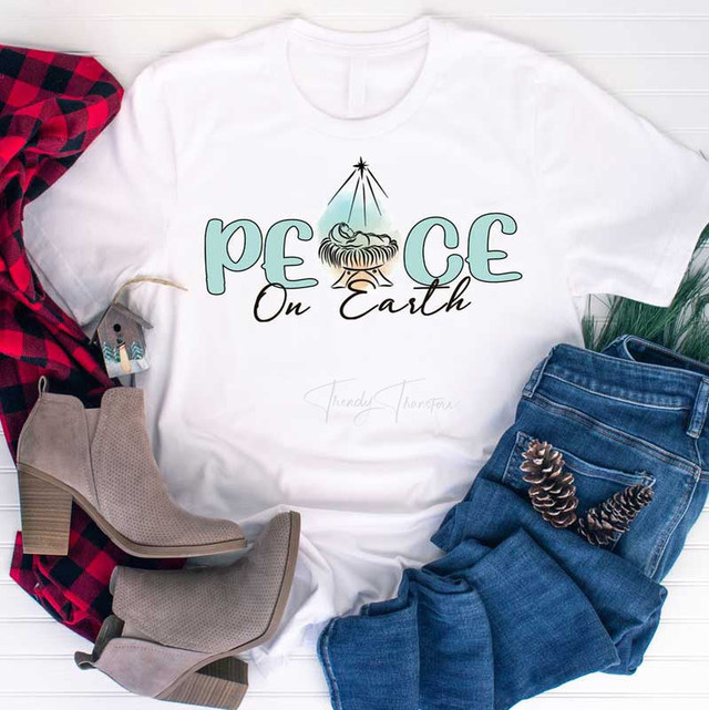 Peace On Earth Sublimation Transfer, white T-shirt with a Christmas-themed design of a nativity scene and the text 'Peace On Earth' in green and black, paired with blue jeans, beige ankle boots, a red and black plaid shirt, and pinecones on a white background.