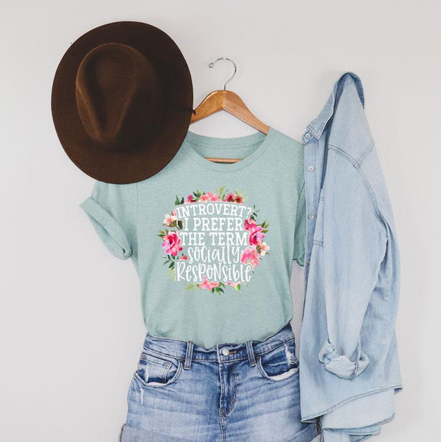 Introvert? I Prefer The Term Socially Responsible Screen Print Transfer, green T-shirt with floral design and humorous message on a hanger with a brown hat and denim jacket.