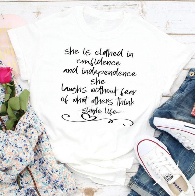 She Is Clothed In Single Life Sublimation Transfer, white T-shirt with inspirational quote about confidence, independence, and laughter in black handwritten style font on a wooden background.