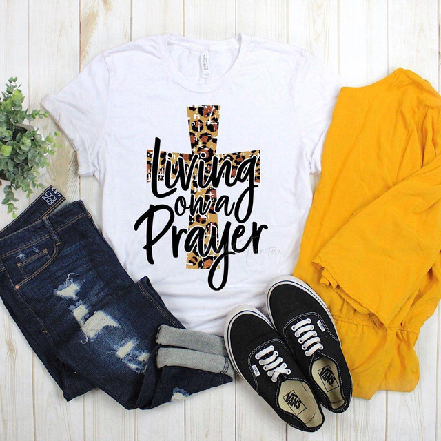 Living On A Prayer with Leopard Cross Sublimation Transfer, product image showing a white t-shirt with a leopard print cross design and the text 'Living On A Prayer', paired with distressed blue jeans, black Vans sneakers, and yellow pants.