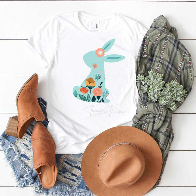 Boho Bunny Sublimation Transfer, graphic of a bunny with floral design on a white T-shirt displayed with denim shorts, brown boots, a hat, a scarf, and a small plant.