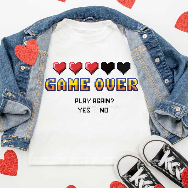 Game Over Sublimation Transfer, an image of a white t-shirt with pixelated hearts and text design, laid flat with a denim jacket and sneakers for display.