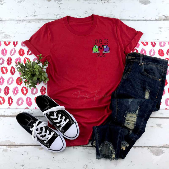 Love Is SUS POCKET/MASK-FINAL STOCK- Screen Print Heat Transfer, red T-shirt with Among Us graphic and text, paired with distressed jeans and black and white sneakers on a white wooden surface with kiss print paper and greenery.
