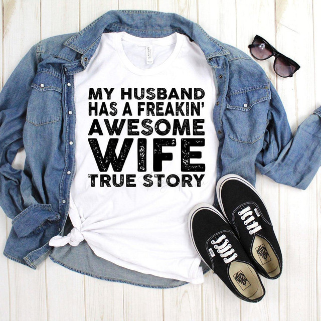 My Husband has a Freakin' Awesome Wife Sublimation Transfer T-shirt, displayed with a denim jacket, sunglasses, and Vans sneakers on a wooden background.