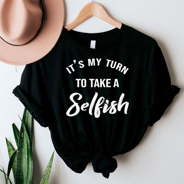 It's My Turn To a Selfish FINAL STOCK Screen Print Heat Transfer, black t-shirt with white text and knot at the bottom, hanging on a wooden hanger with a hat and green plant nearby.
