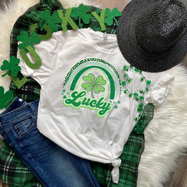 Lucky Rainbow Sublimation Transfer, white t-shirt with a green leprechaun hat and rainbow design, St. Patrick's Day themed accessories including shamrock necklace and green foam shamrocks, and a black sun hat, decorated with green plaid fabric and shamrock-themed decorations.