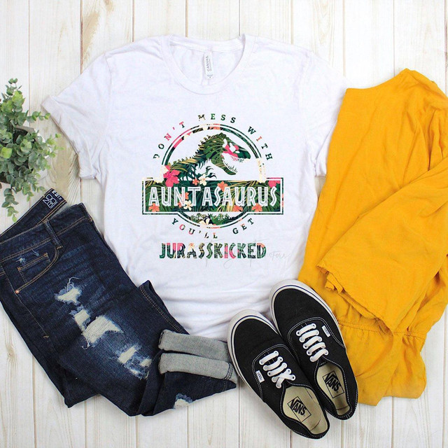 Don't Mess With Auntasaurus You'll Get Jurasskicked Tropical Sublimation Transfer, white T-shirt with colorful dinosaur-themed graphic and text, paired with distressed jeans, black Vans sneakers, and a yellow jacket on a light wooden surface.