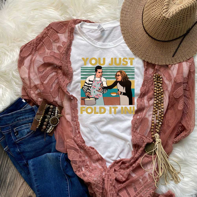 You Just Fold It In Sublimation Transfer, white t-shirt with a graphic design of people cooking and the text "YOU JUST FOLD IT IN!", surrounded by a pink lace shawl, a straw hat, denim jeans, and accessories, laid out on a fluffy white surface.