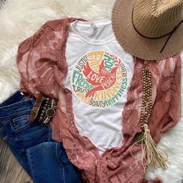Hope Peace Love Dove Sublimation Transfer, colorful design with words like hope, peace, love in a circular pattern on a white t-shirt, placed among jeans, a straw hat, lace cover-up, bracelets, and accessories on a soft surface.