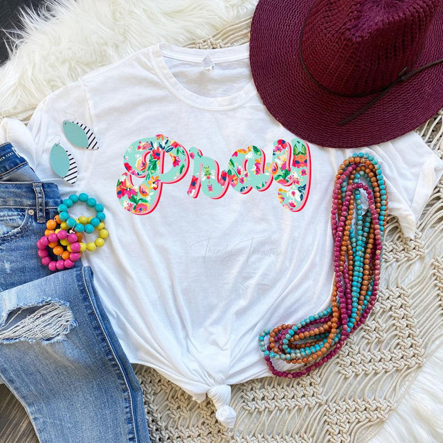 Pray Sublimation Transfer-1655991809, colorful floral text design on a white t-shirt, surrounded by accessories including a red hat, beaded necklaces, and earrings.