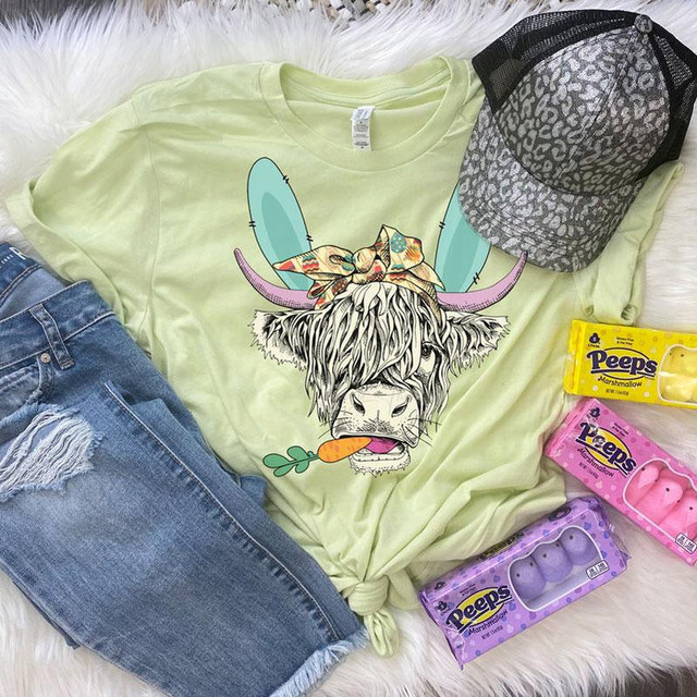 Shaggy Cow Easter Screen Print Heat Transfer, graphic of a cheerful cow with shaggy hair, wearing a colorful bow and bunny ears on a pastel yellow T-shirt.