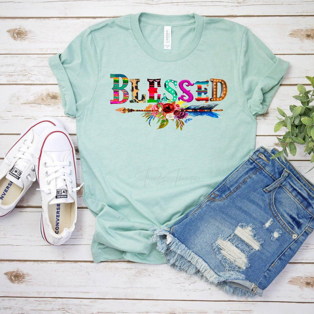 Blessed Serape and Leopard Screen Print Heat Transfer, colorful and stylish design on a light green t-shirt, paired with distressed denim shorts and white Converse sneakers, displayed on a white wooden background with a green plant.