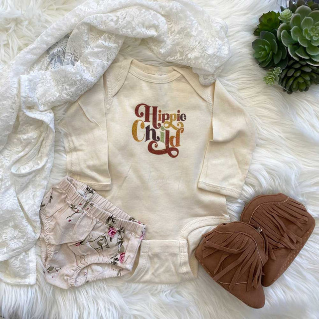 Hippie Child POCKET/INFANT Screen Print Heat Transfer, baby clothes set including a cream infant onesie with a 'Hippie Child' colorful screen print, floral print shorts, cream lace headband, and brown fringed boots on a white furry background with green succulents.