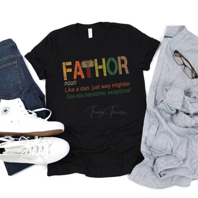FaThor Screen Print Heat Transfer, black T-shirt with colorful print design of the word 'FaThor' and humorous text, paired with denim jeans, white sneakers, gray shirt with glasses, and casual accessories, laid out on a white background.