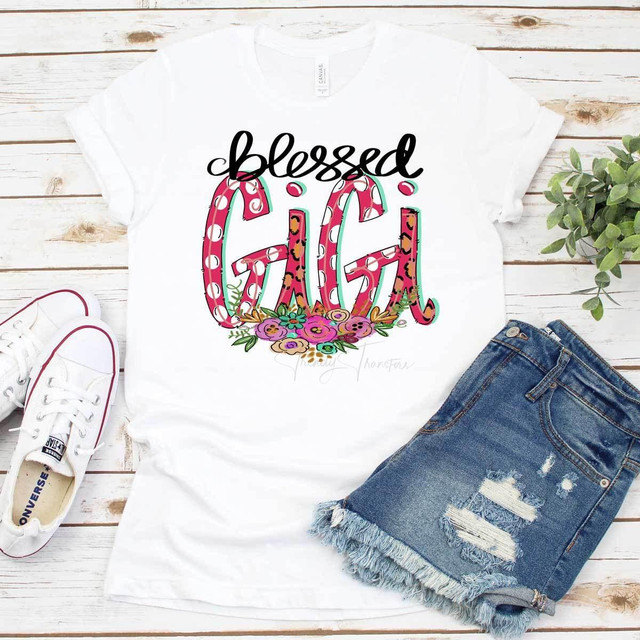 Blessed GIGI Sublimation Transfer T-shirt with colorful floral and animal print design, wide neckline on white fabric, paired with distressed denim shorts and white sneakers, on a wooden surface with green plant for a trendy, casual look.
