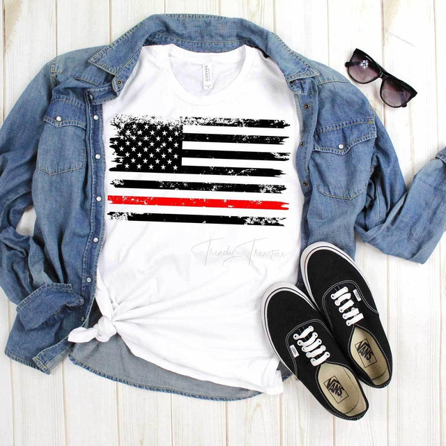 Flag With Thin Red Line Sublimation Transfer, a graphic T-shirt featuring a black and red American flag design with a distressed style, laid on a wooden surface with a denim jacket, sunglasses, and Vans shoes.